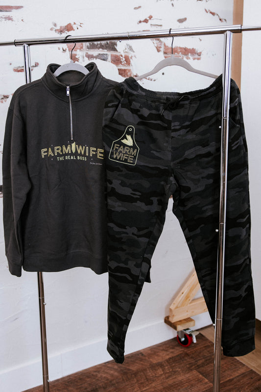 Black 'Farm Wife' sweatshirt and camouflage pants on a rack against a white wall.