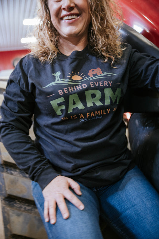 Person wearing a black sweatshirt with a farm-themed design, sitting in front of a red vehicle.