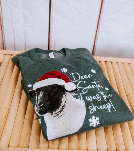 Green t-shirt with a sheep wearing a Santa hat and text on a wooden surface.