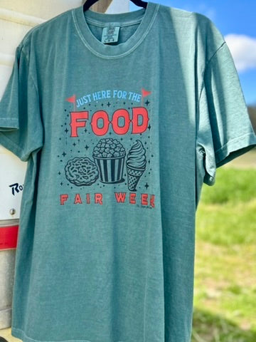 Teal t-shirt with food-themed graphic and text on a hanger against a blurred outdoor background