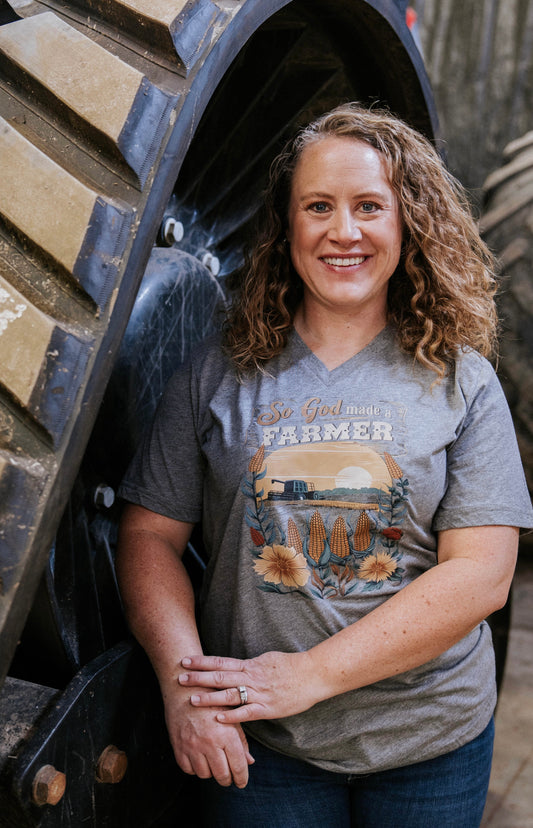 Woman wearing a 'So God Made a Farmer' t-shirt standing next to a large agricultural tire.