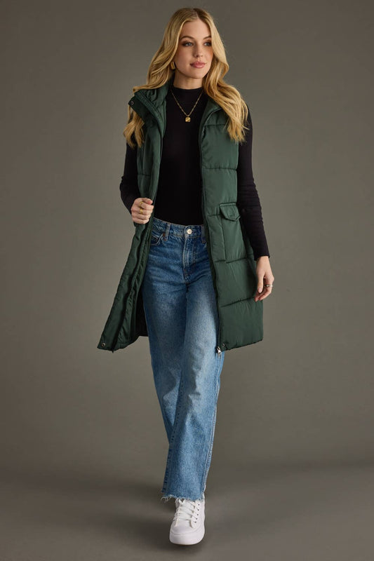 Woman wearing a green puffer vest, black top, blue jeans, and white sneakers on a gray background
