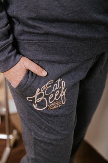 Dark gray sweatpants with 'Eat Local Support Farmers' text on a blurred indoor background