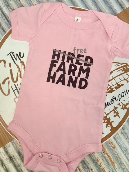 Pink baby onesie with 'free hired farm hand' text on a wooden surface.