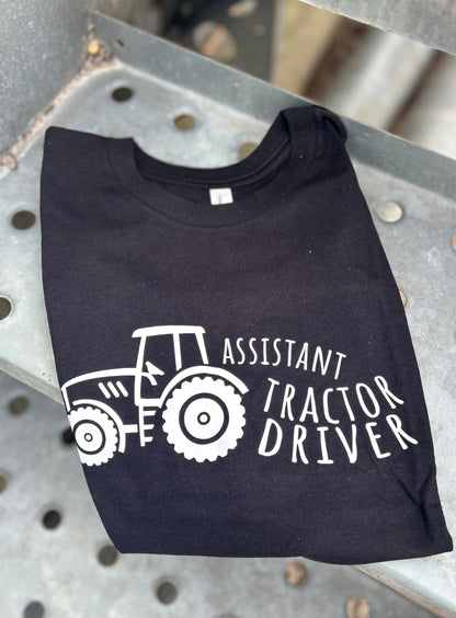 Black t-shirt with white tractor graphic and text 'Assistant Tractor Driver' on a metal surface.