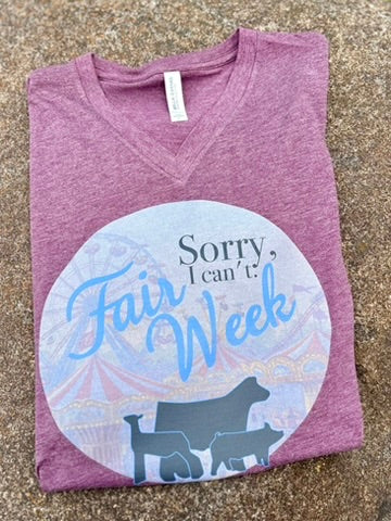 Folded maroon t-shirt with 'Sorry, I can't. Fair Week' graphic on a textured surface