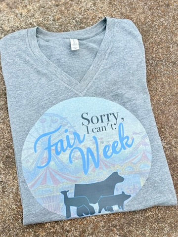 Gray V-neck shirt with 'Sorry, I can't. Fair Week' text and animal silhouettes on a textured surface.