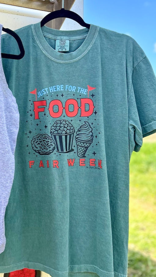 Green t-shirt with red and black text and graphics on a hanger against a natural background
