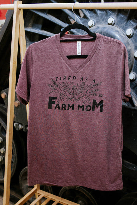 Maroon t-shirt with 'Tired as a Farm Mom' text on a wooden rack with large wheels in the background