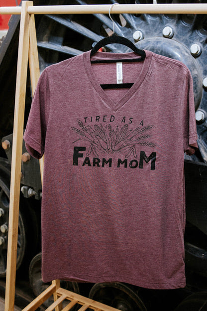 Maroon t-shirt with 'Tired as a Farm Mom' text on a wooden rack with large wheels in the background