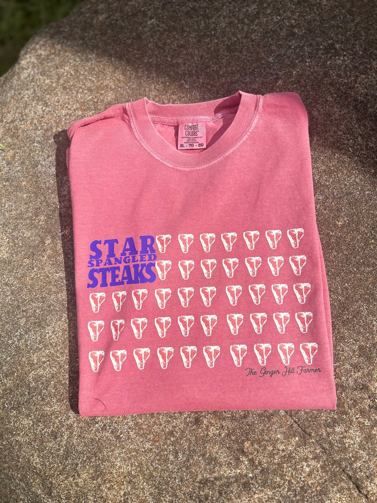 Pink t-shirt with 'Star Spangled Steaks' text and graphics on a concrete surface