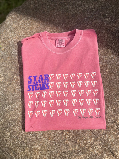 Pink t-shirt with 'Star Spangled Steaks' text and graphics on a concrete surface