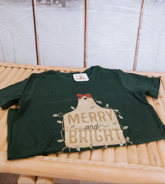 Green t-shirt with 'Merry and Bright' design on a wooden surface with a snowy window background