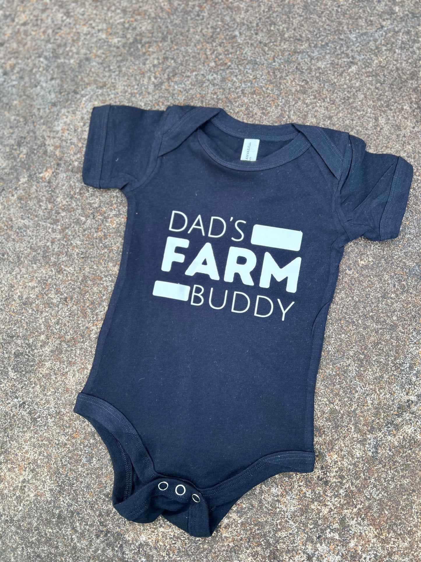 black baby onesie with 'Dad's Farm Buddy' text on a concrete surface