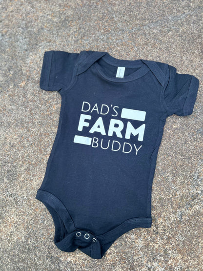 black baby onesie with 'Dad's Farm Buddy' text on a concrete surface