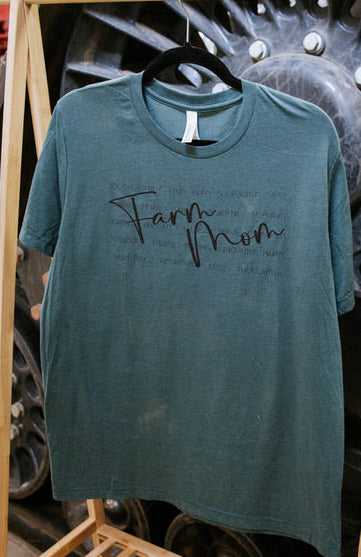 Green t-shirt with 'Farm Mom' text on a wooden rack in front of large machinery.