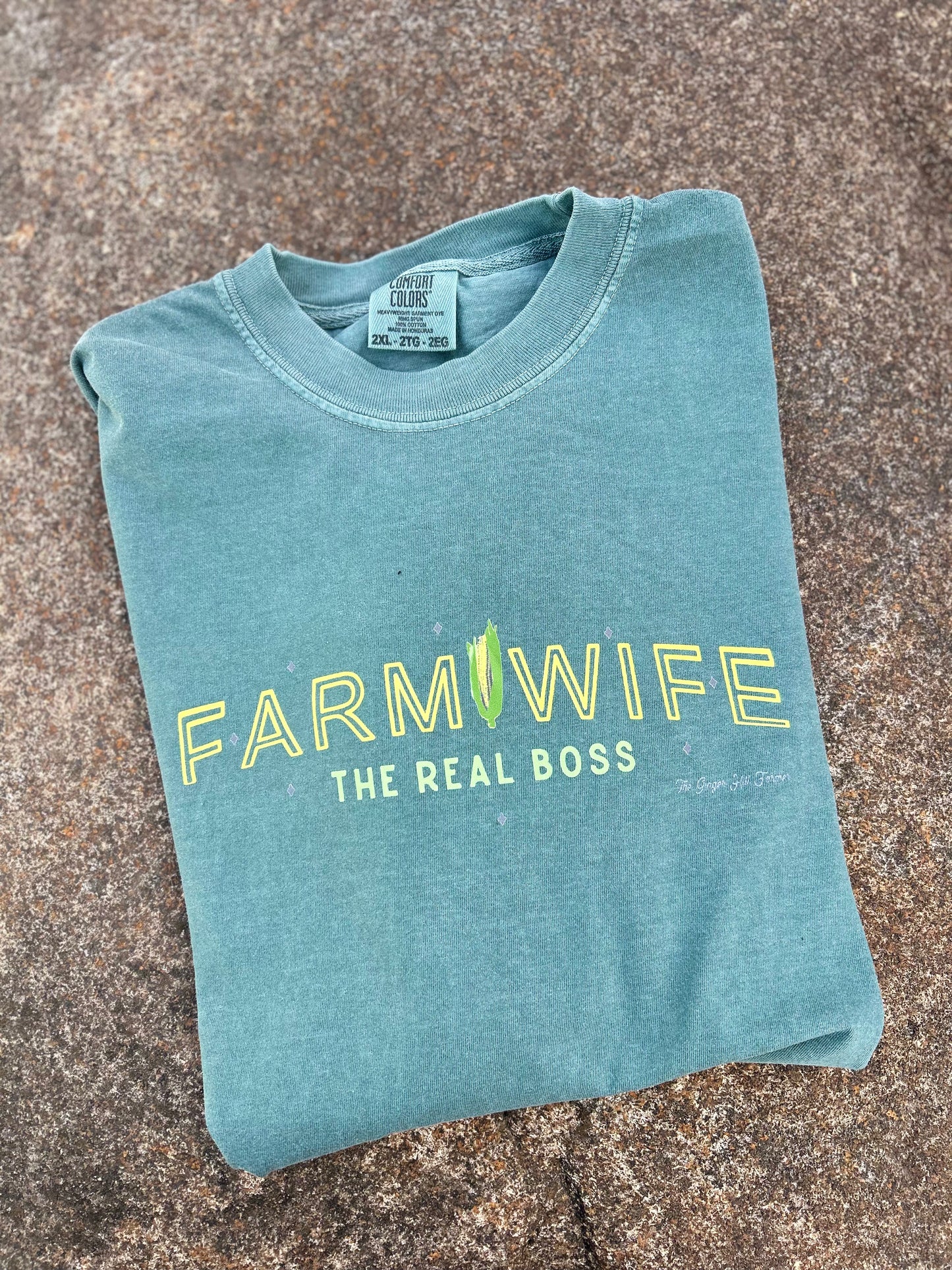 Folded teal t-shirt with 'Farmwife, The Real Boss' text on a textured surface