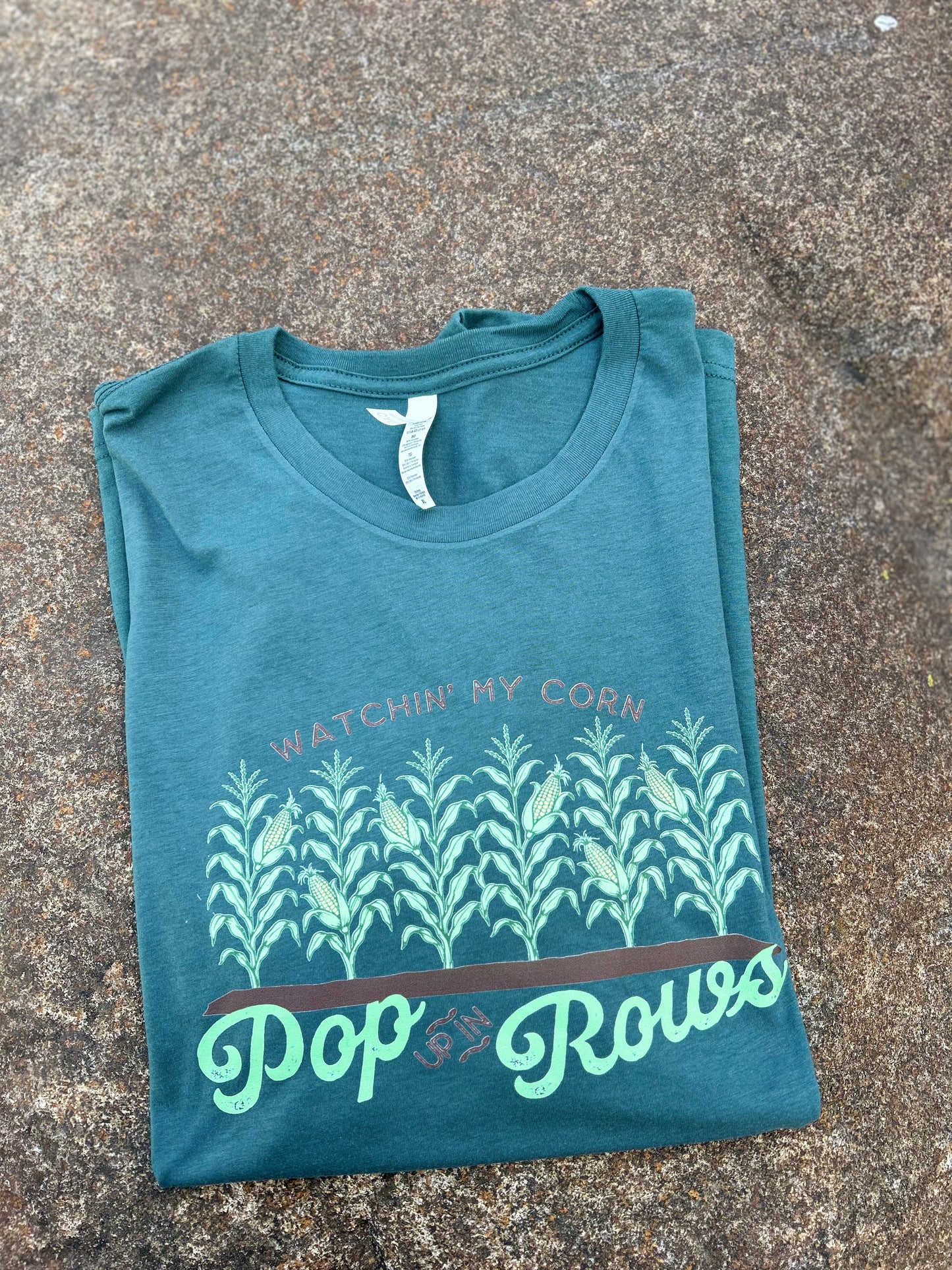 Folded teal t-shirt with corn plant design and text on a concrete surface