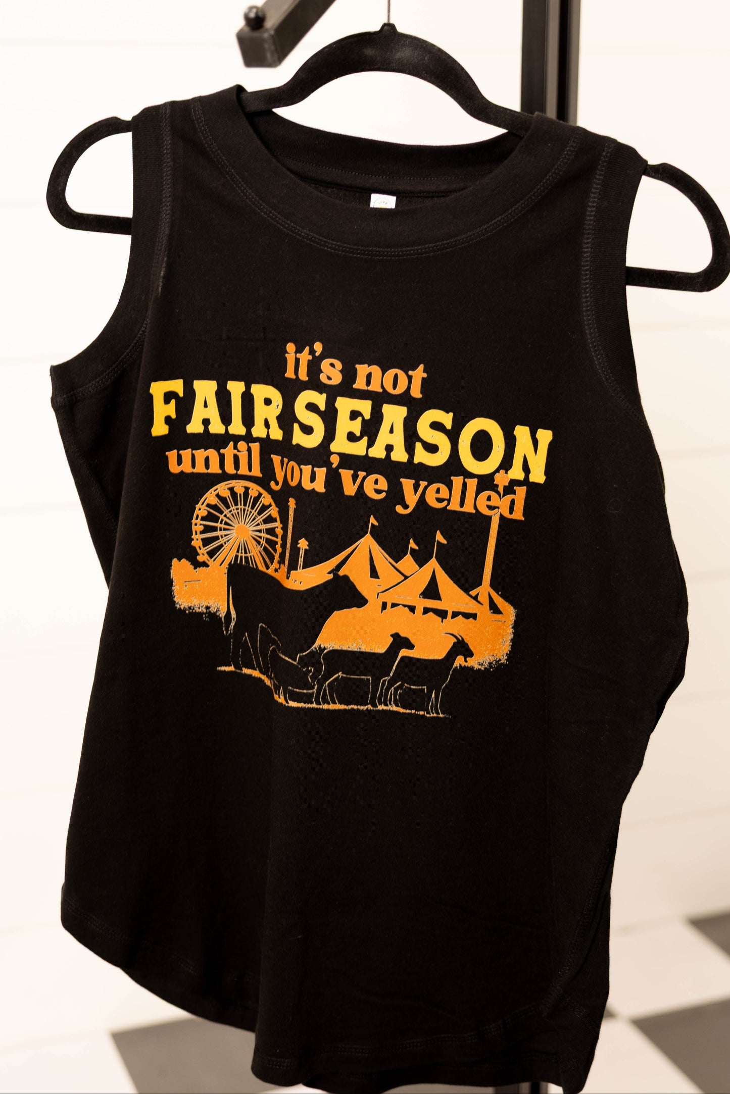 Black tank top with yellow text and graphic on a hanger against a checkered floor background
