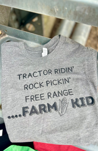 Gray t-shirt with 'Tractor Ridin' Rock Pickin' Free Range Farm Kid' text on a metal surface.