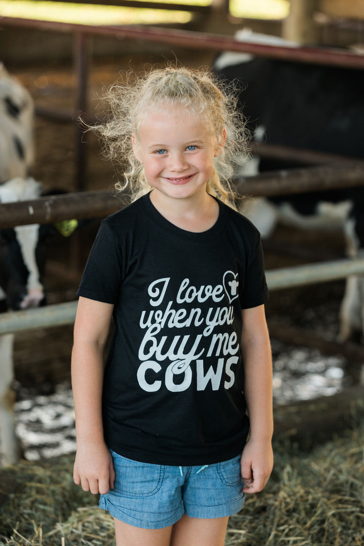 Child wearing a black t-shirt with text standing in a barn with cows in the background