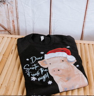 Black t-shirt with a pig wearing a Santa hat and text, placed on a wooden surface with a snowy window background.