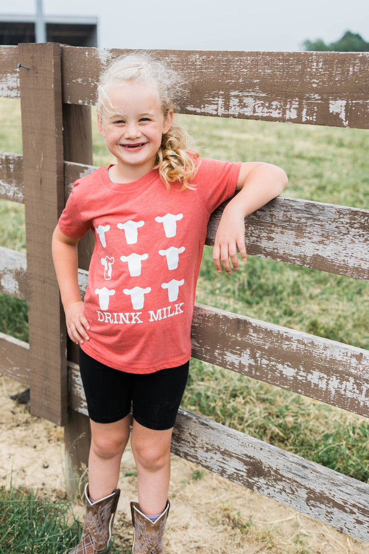 Child wearing a red shirt with cow graphics and 'Drink Milk' text, standing by a wooden fence on a farm.
