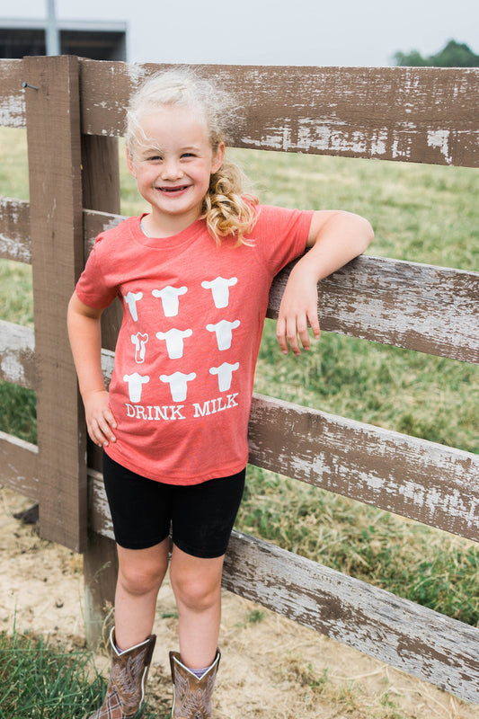 Child wearing a red shirt with cow graphics and 'Drink Milk' text, standing by a wooden fence on a farm.
