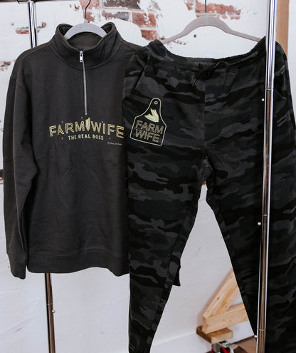 Black 'Farm Wife' sweatshirt and camouflage pants on a rack against a white wall.